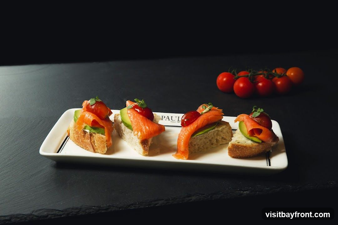Four small open-faced sandwiches on a white rectangular plate. Each sandwich has a slice of bread topped with avocado and a piece of smoked salmon, garnished with a small red sauce dollop and microgreens. A cluster of cherry tomatoes is visible in the background. The plate has a subtle logo that reads "PAUL depuis 1889".