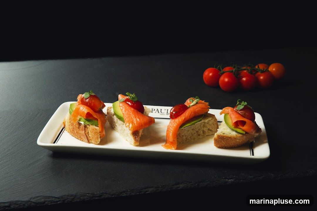 Four small open-faced sandwiches on a white rectangular plate. Each sandwich has a slice of bread topped with avocado and a piece of smoked salmon, garnished with a small red sauce dollop and microgreens. A cluster of cherry tomatoes is visible in the background. The plate has a subtle logo that reads "PAUL depuis 1889".