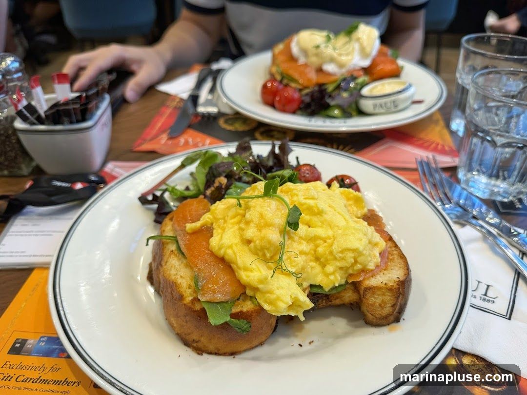 A close-up shot of two plates of brunch at a restaurant. The plate in the foreground features scrambled eggs and smoked salmon on toast, garnished with microgreens and cherry tomatoes. The plate in the background shows eggs Benedict with smoked salmon and hollandaise sauce, also with cherry tomatoes and a side salad. The restaurant's branding,