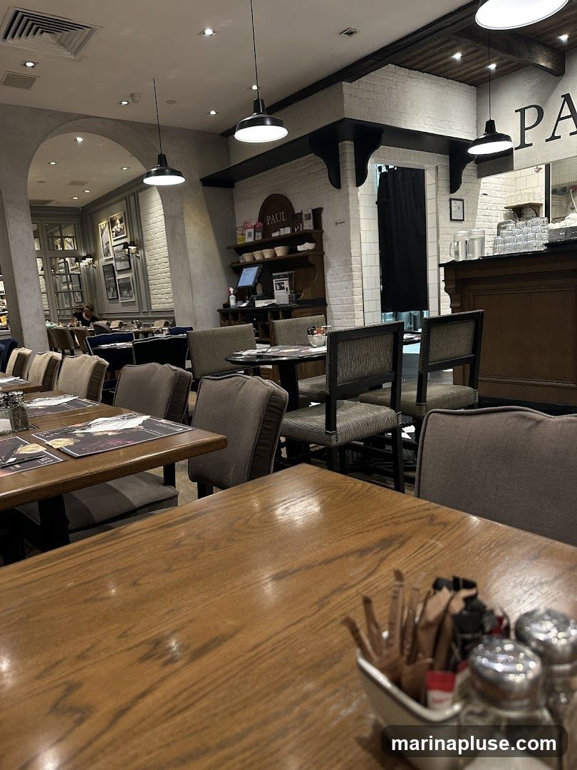 The image shows the interior of a restaurant with wooden tables and chairs. There are several tables set with placemats and cutlery. In the background, there is a counter area with a display shelf that has the word "PAUL" on it. The walls are decorated with framed pictures and white brickwork. Pendant lights hang from the ceiling.