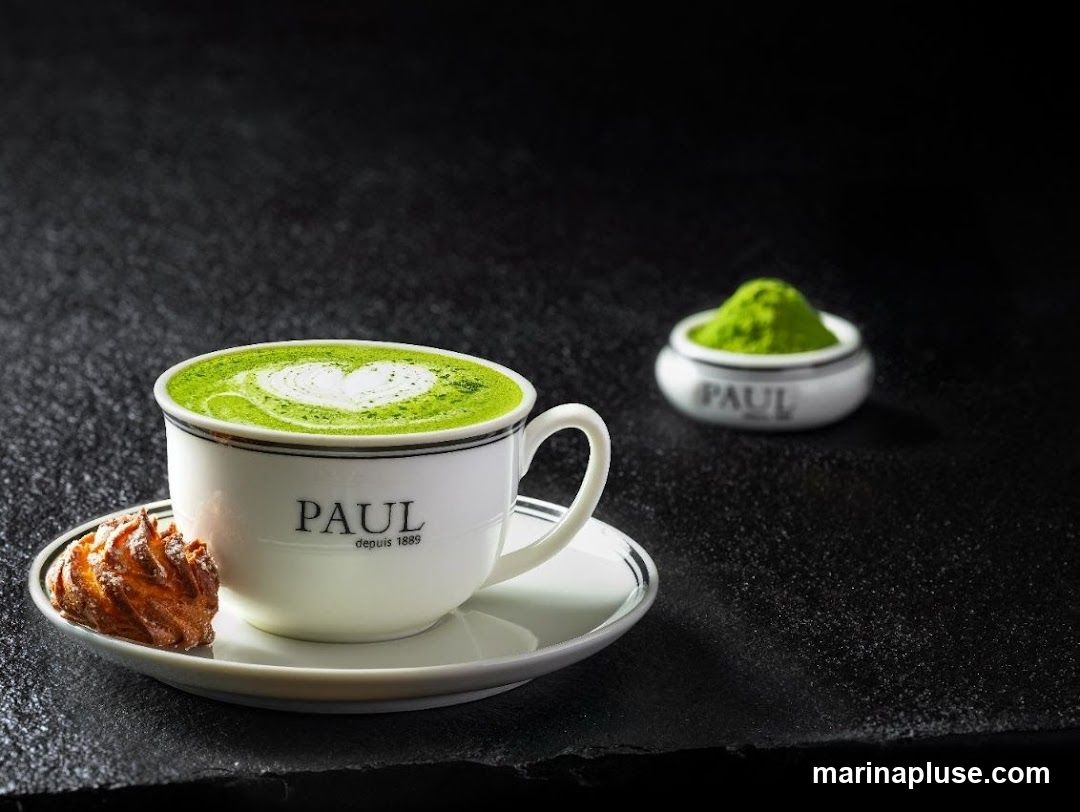 A close-up shot of a matcha latte in a white cup with a heart design on top, placed on a saucer with a small pastry. A small bowl of matcha powder is visible in the background. The cup and bowl have the PAUL logo.