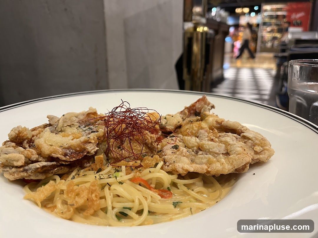 A close-up shot of a plate of pasta topped with fried soft shell crab. The pasta appears to be a creamy sauce with herbs and some red pepper flakes. The soft shell crab is golden brown and crispy, garnished with thin red threads. In the background, a blurred image of a restaurant interior with checkered flooring and a person walking in the distance. A glass of water is visible on the right side of the frame.