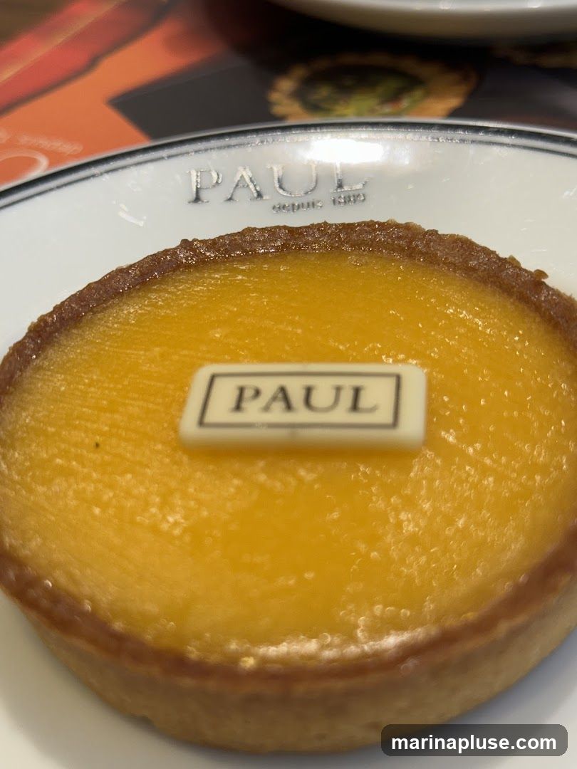 A close-up shot of a lemon tart from PAUL bakery. The tart has a golden crust and a bright yellow filling. A small white rectangular piece of chocolate with the PAUL logo is placed on top of the tart. The plate has the PAUL logo embossed on it.