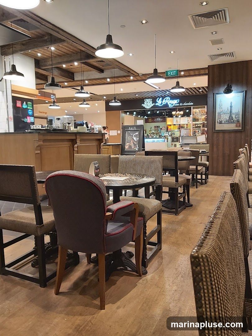 The image shows the interior of a cafe with wooden floors and exposed wooden beams on the ceiling. Several tables and chairs are arranged for seating. In the background, a counter area is visible with a sign that reads "Le Rouge Cuisine & Chocolates". There are also shelves stocked with products and a framed picture of the Eiffel Tower on the wall.