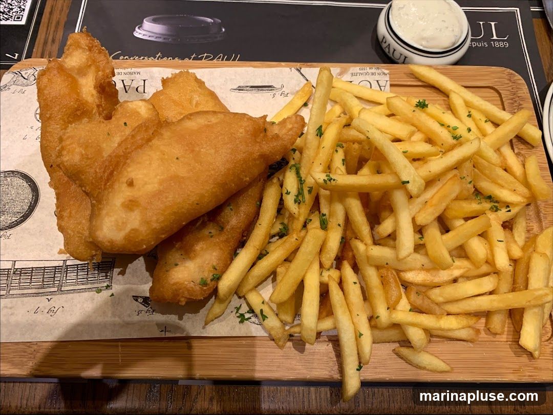 A wooden tray holds a serving of battered fish pieces and a large portion of french fries, garnished with chopped parsley. A small ramekin of white sauce is visible in the upper right corner. The tray is placed on a table with a placemat that has text and illustrations. The brand name "PAUL" is visible on the ramekin and the placemat.