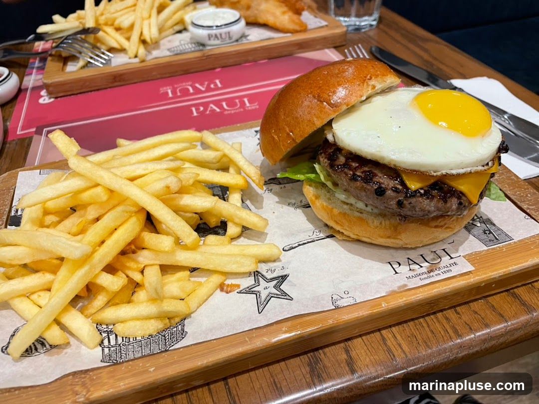 A close-up shot of a burger with a fried egg and cheese, served with a side of french fries. The food is presented on wooden boards lined with paper featuring the PAUL logo.