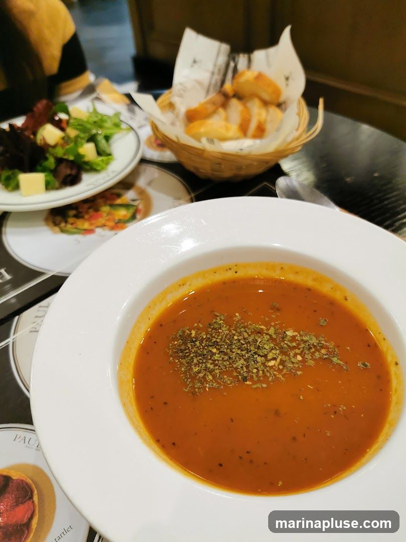 A bowl of tomato soup topped with herbs sits on a table next to a salad with cheese and greens, and a basket of sliced bread. A menu with the PAUL logo is visible under the soup bowl.