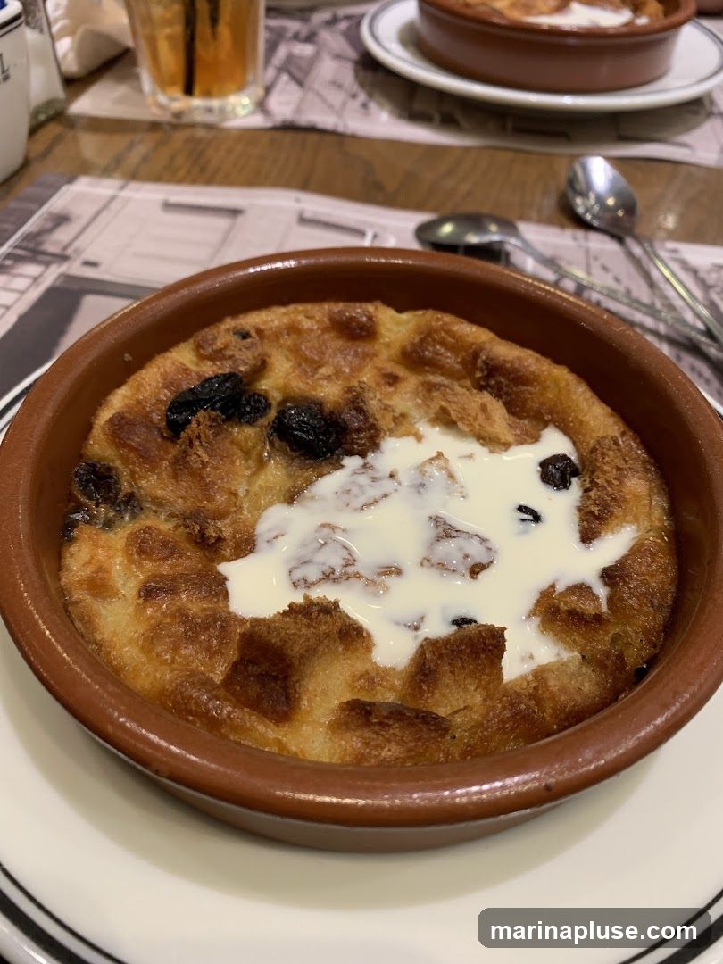 A close-up shot of a bread pudding served in a brown ceramic dish. The pudding is golden brown and appears to be made with bread, raisins, and topped with a dollop of cream. The dish is placed on a white plate with black trim. In the background, a glass of amber liquid and cutlery are visible on a table with a patterned placemat.