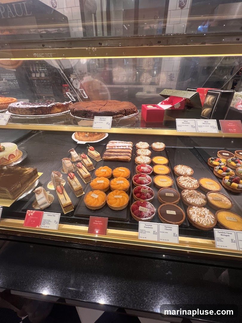 A display case filled with various pastries and cakes. Visible items include a large chocolate cake, several fruit tarts, eclairs, and smaller individual tarts. Some items have price labels.