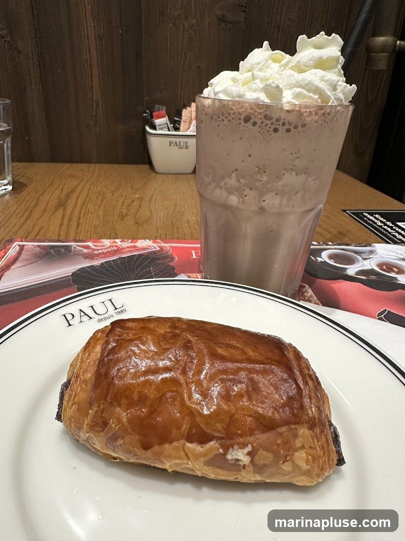 A chocolate croissant sits on a white plate with the PAUL logo. Next to it is a tall glass filled with a chocolate frappe topped with whipped cream and a straw. In the background, a wooden wall and a small container with PAUL branding holding sugar packets and stirrers are visible.