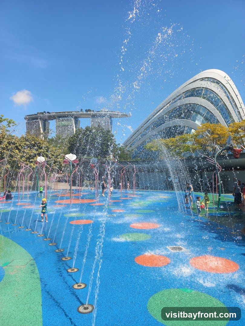 A vibrant water playground with children playing amidst spraying fountains under a clear blue sky. In the background, the iconic Marina Bay Sands hotel and a modern, curved glass building are visible, suggesting an urban setting. The ground of the playground is blue with colorful circular patterns and green accents.