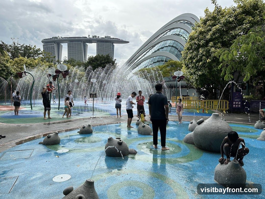 A group of people, including children and adults, are enjoying a water play area. Water jets are spraying from various structures, including flower-shaped fountains and animal-shaped sculptures. In the background, the distinctive architecture of Marina Bay Sands and Gardens by the Bay can be seen. The ground is wet and colorful, with blue, green, and white patterns.