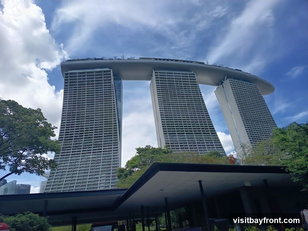 A low-angle shot of the Marina Bay Sands hotel in Singapore, with its distinctive three towers connected by a large, boat-shaped structure at the top. The sky is blue with scattered white clouds. Lush green trees frame the bottom of the image, and a covered walkway or pavilion is visible in the foreground.
