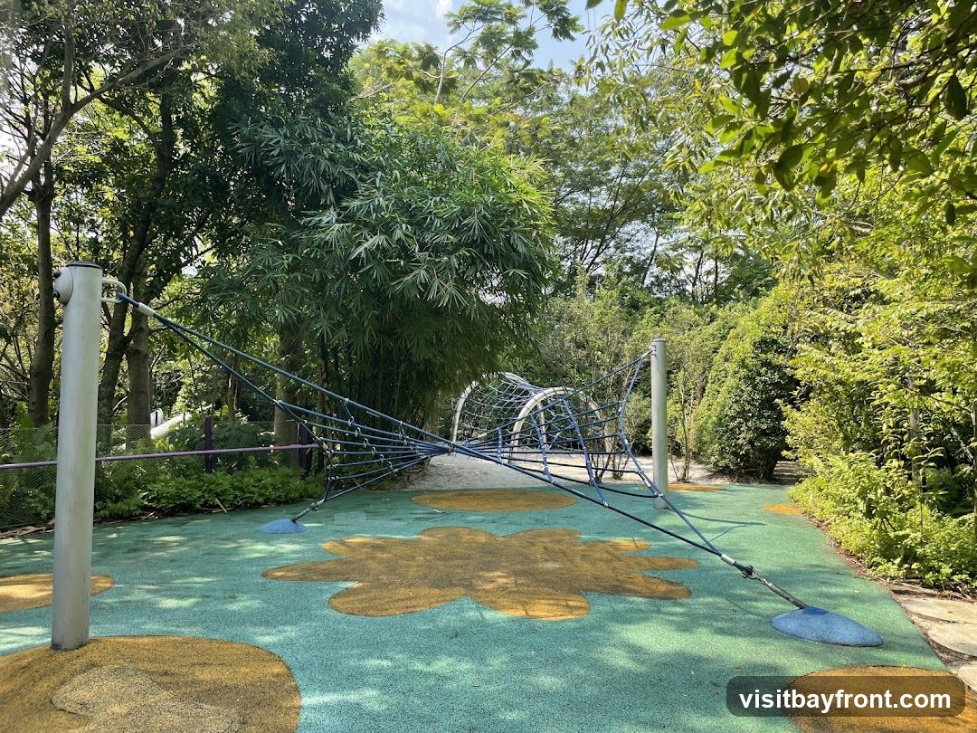 A wide shot of a playground with a large rope climbing structure in the center. The ground is covered in a green and yellow rubberized surface with flower-like patterns. Tall trees and lush greenery surround the playground, creating a natural and shaded environment. The sky is visible through the leaves, suggesting a bright, sunny day.
