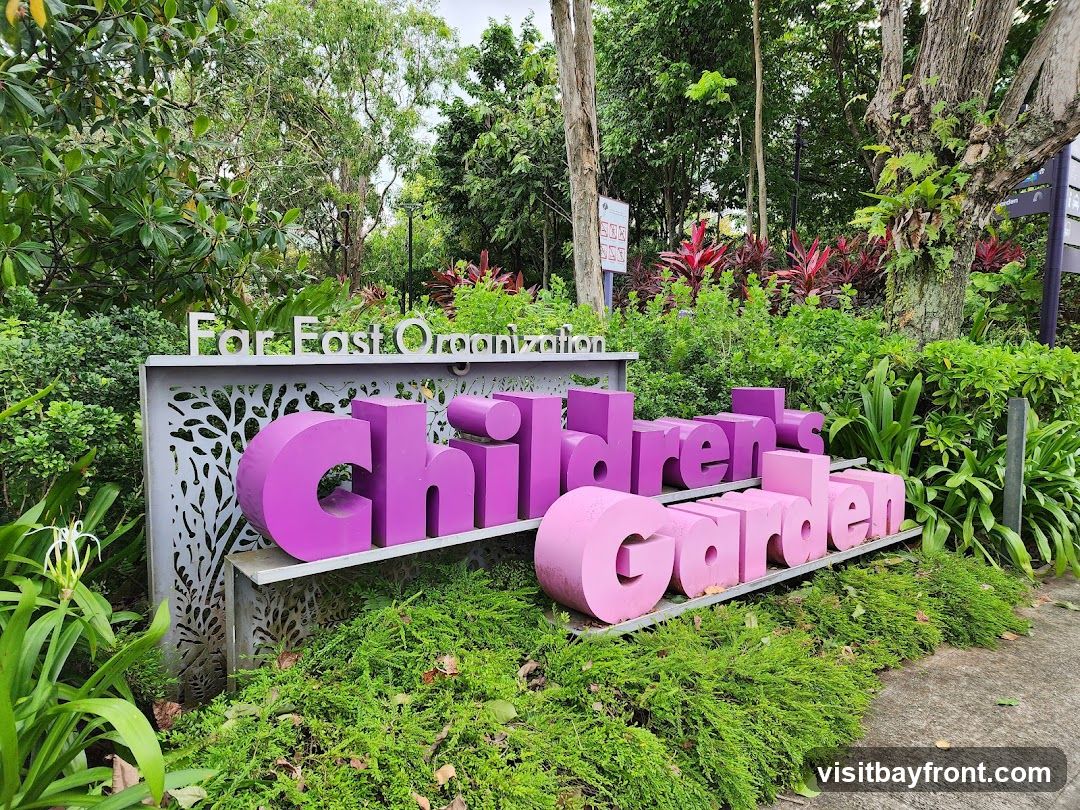 A sign for the Children's Garden, sponsored by Far East Organization, is displayed outdoors amidst lush greenery. The sign features large, three-dimensional purple and pink letters spelling out "Children's Garden." Above the letters, the words "For East Organization" are visible in silver lettering on a decorative metal panel. The surrounding area is filled with various plants, trees, and foliage, suggesting a park or garden setting.