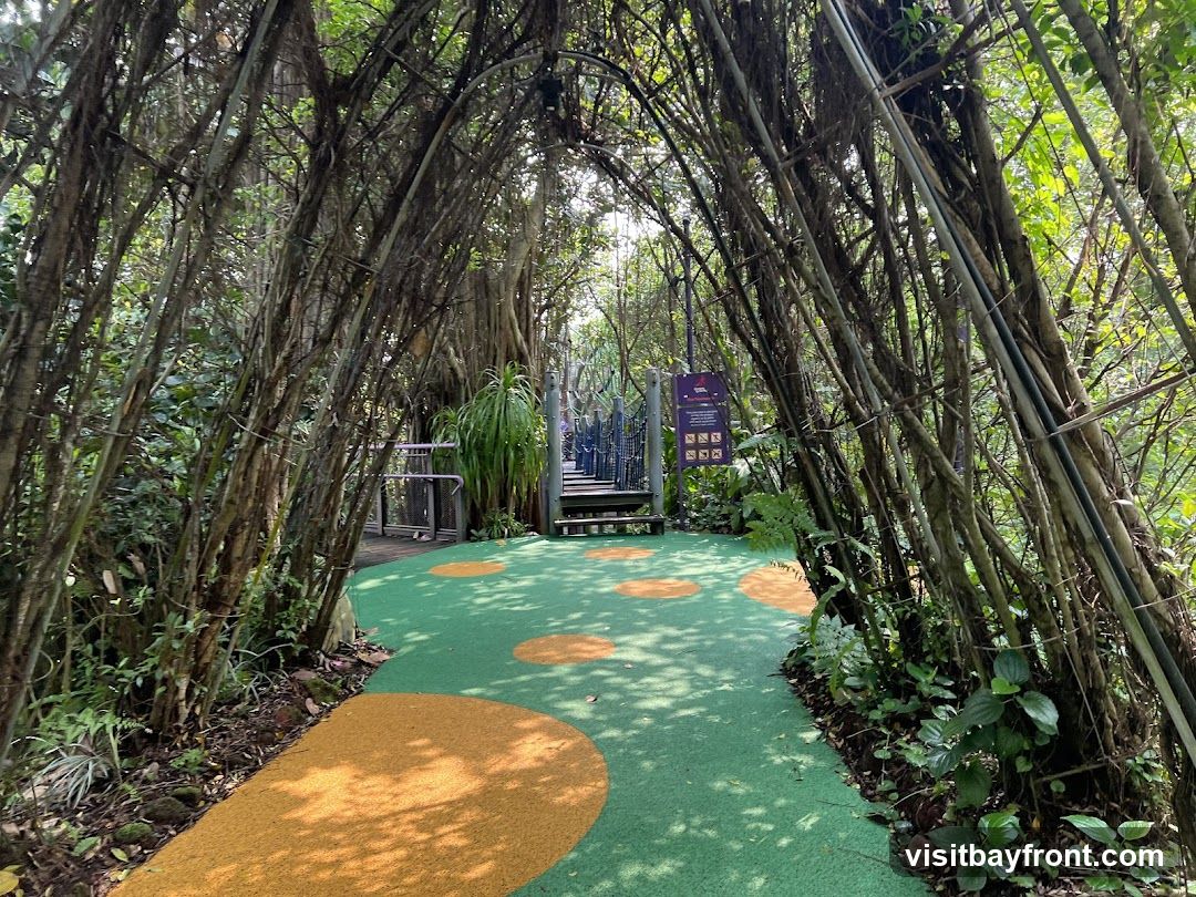 A winding path through a dense forest, with trees forming an archway overhead. The ground is covered in green artificial turf with orange circular patterns. In the background, a wooden walkway and a purple sign with text and icons are visible.
