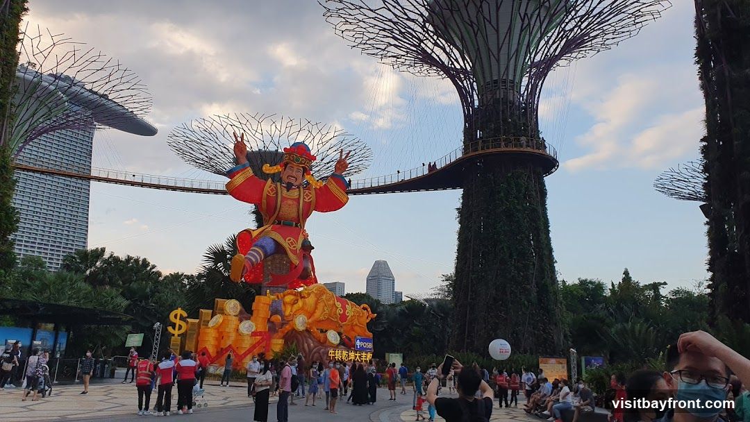A large, colorful lantern display featuring a figure in traditional Chinese attire, seated on a bull and surrounded by gold coins, is set up in an outdoor area. In the background, the iconic Supertrees of Gardens by the Bay are visible, along with a skywalk connecting them. A tall modern building stands in the distance. Many people are gathered, observing the display and taking photos. The sky is partly cloudy.