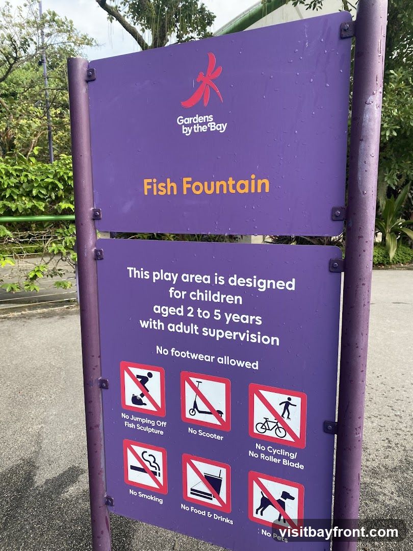 A purple sign with the Gardens by the Bay logo and the text "Fish Fountain". Below this, it states that the play area is designed for children aged 2 to 5 years with adult supervision and that no footwear is allowed. The sign also lists several prohibited activities with corresponding icons: no jumping off fish sculpture, no scooter, no cycling/roller blade, no smoking, no food & drinks, and no pets.