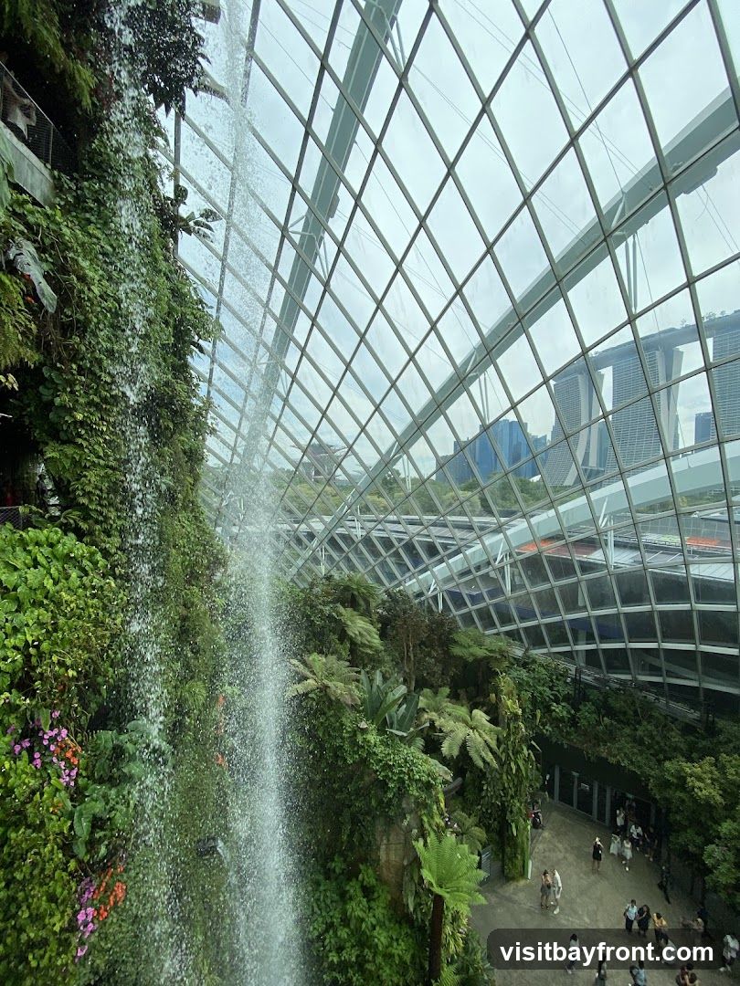 A lush indoor garden with a tall waterfall cascading down a moss-covered rock face. The greenhouse structure features a diamond-patterned glass ceiling, allowing natural light to filter through. In the background, modern skyscrapers are visible through the glass. People are seen walking on a pathway below the greenery.