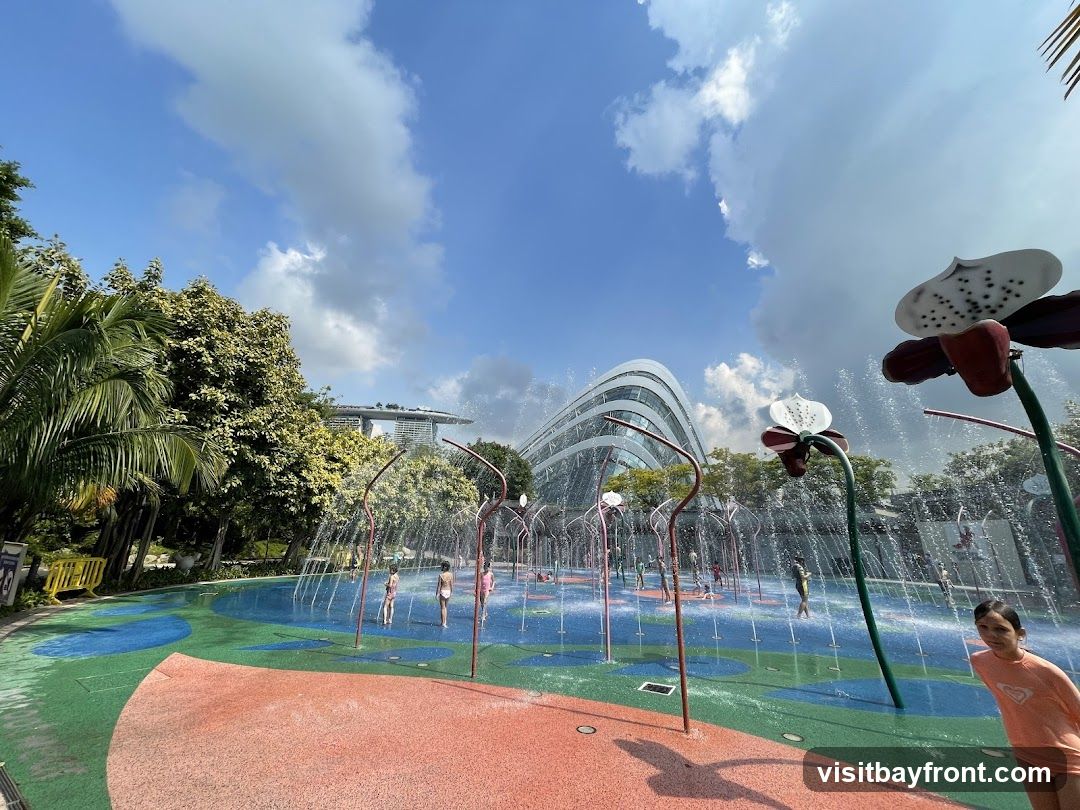 A vibrant water playground with children playing amidst spraying water features shaped like flowers. In the background, a modern, curved glass building stands against a bright blue sky with fluffy clouds. Lush green trees surround the area, creating a lively and refreshing atmosphere.