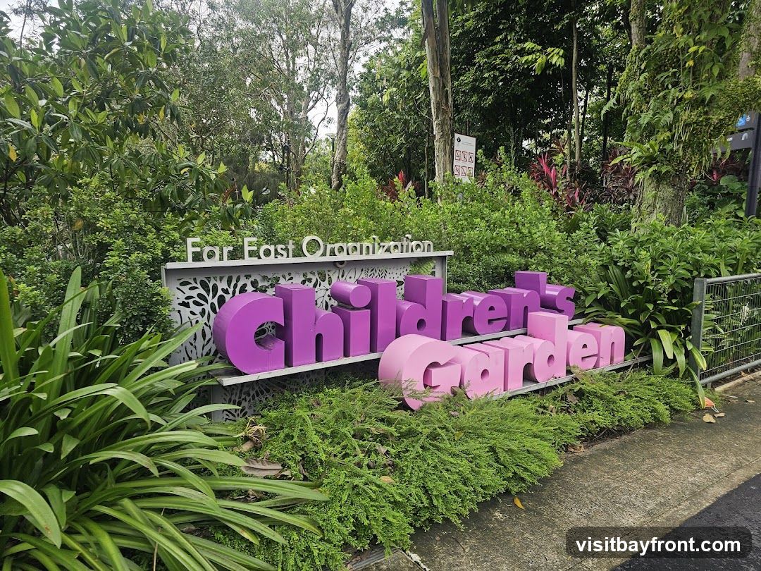 A sign for the Children's Garden, sponsored by Far East Organization, is set against a backdrop of lush greenery. The sign features large, three-dimensional letters in shades of purple and pink, spelling out "Children's Garden." Above the colorful letters, the words "Far East Organization" are displayed in white lettering on a grey panel with a decorative cutout pattern. The sign is surrounded by various plants and foliage, with a concrete path visible in the foreground.