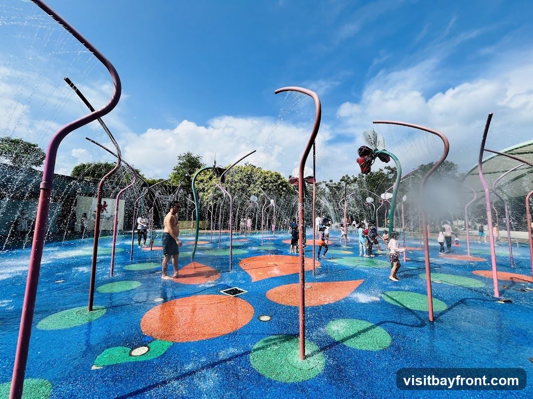 A vibrant water play area with numerous spray features and colorful ground markings. Children and adults are seen enjoying the water under a bright blue sky with scattered clouds. The ground is blue with large orange and green circular and oval shapes. Metal structures with water jets arch over the play area, some resembling flowers.