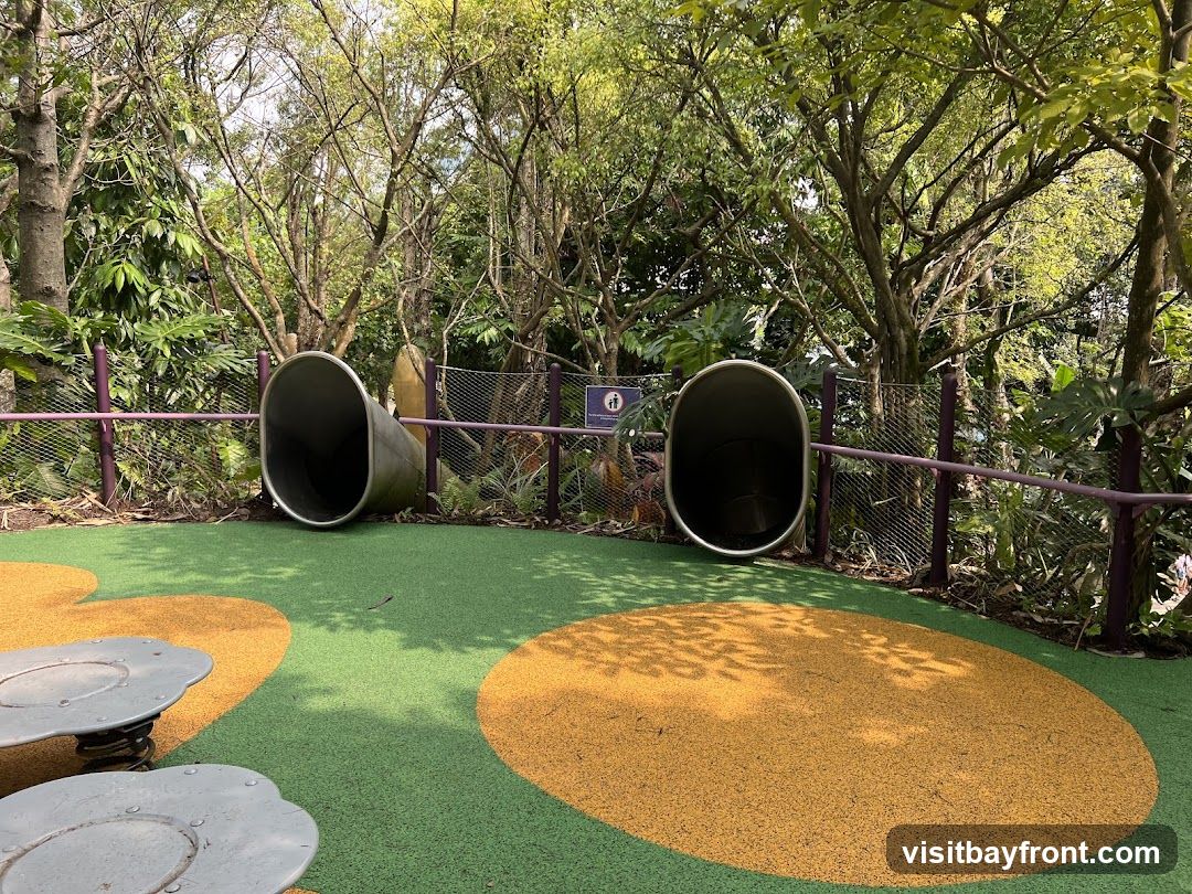 A playground area with two large metal tube slides. The ground is covered in green and yellow rubber surfacing with large circular patterns. In the foreground, there are two spring-mounted play structures. The area is surrounded by lush green trees and foliage. A small sign with a pictogram is visible on a fence in the background.