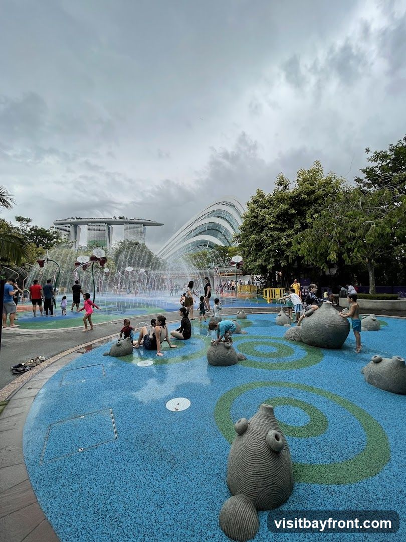 A lively water playground with children and adults enjoying the splash pads and water features. The ground is a vibrant blue with green spiral designs and whimsical stone sculptures. In the background, the iconic Marina Bay Sands hotel and the distinctive Supertree Grove structures of Gardens by the Bay are visible under a cloudy sky.