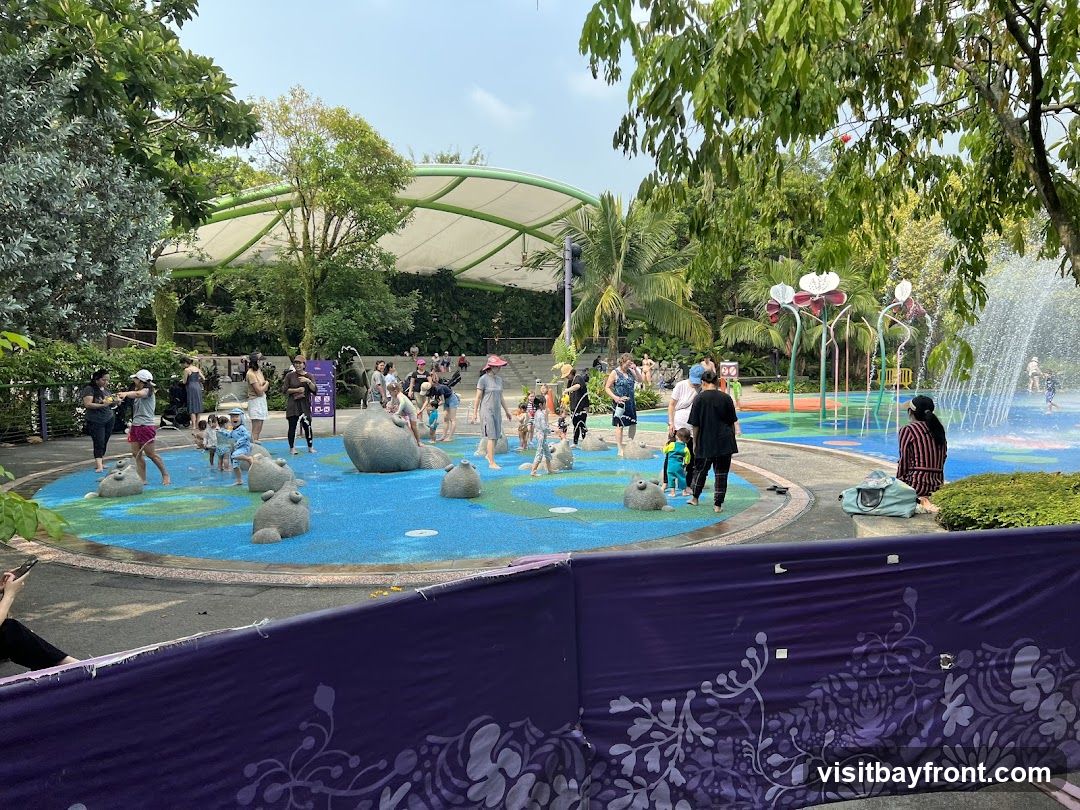 A lively outdoor water play area with children and adults enjoying the sprinklers and water features. The area has a colorful, cushioned ground with various animal-shaped structures. A large, white canopy provides shade over a seating area in the background. Lush greenery surrounds the play zone.