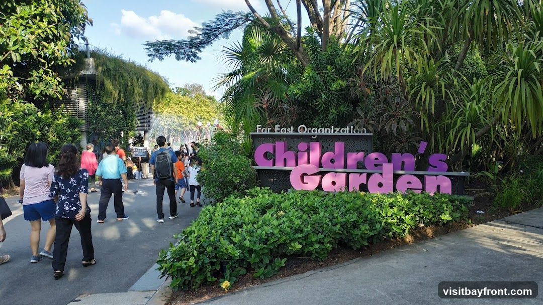 A sign that reads "Far East Organization Children's Garden" is prominently displayed amidst lush greenery. People are walking along a paved path towards the garden entrance, with a building covered in vines visible in the background. The scene suggests a family-friendly outdoor attraction.