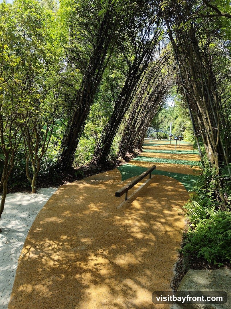 A sun-dappled pathway winds through a lush green park. Trees arch overhead, forming a natural tunnel. The path is made of yellow gravel with green sections. A wooden balance beam is visible on the path. In the distance, playground equipment can be seen.