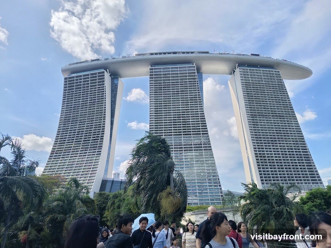 A wide shot of the Marina Bay Sands hotel in Singapore, with its distinctive three towers connected by a large rooftop deck. The hotel is set against a bright blue sky with scattered clouds. In the foreground, a crowd of people are walking, with lush green palm trees and other foliage framing the lower part of the image.