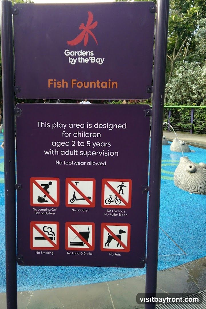 A sign at the Gardens by the Bay Fish Fountain play area lists rules for children aged 2 to 5 years. It states that adult supervision is required and no footwear is allowed. Prohibited activities include jumping off fish sculptures, using scooters, cycling, rollerblading, smoking, bringing food and drinks, and bringing pets. The play area itself, with water features, is visible in the background.
