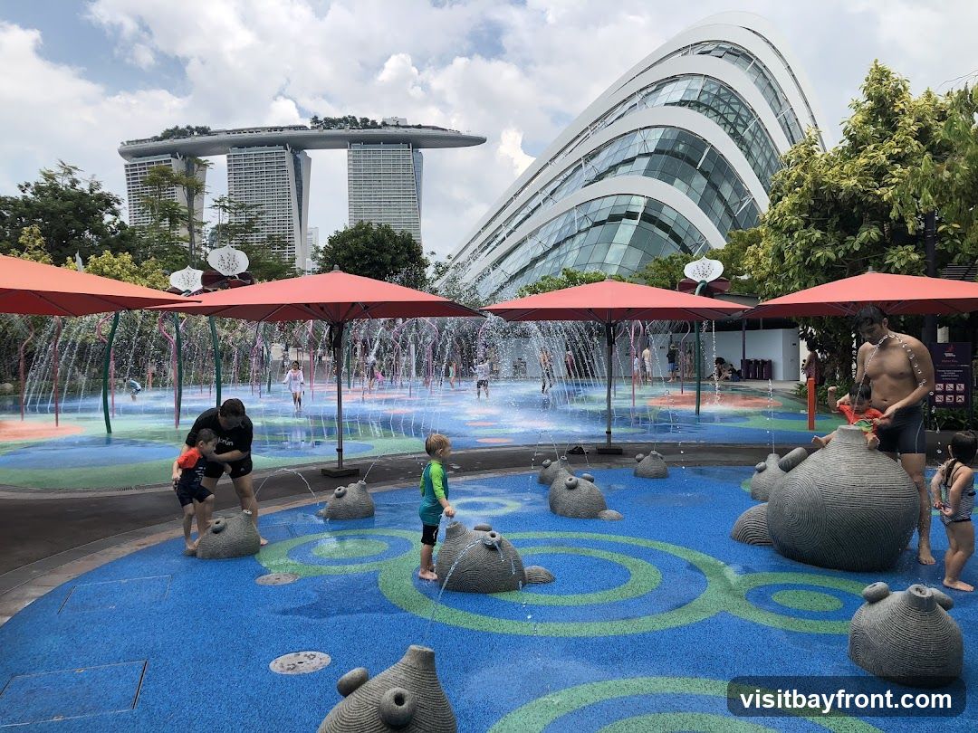 A vibrant water play area with children and adults enjoying the sprinklers and water features. Red umbrellas provide shade, and whimsical stone sculptures are scattered throughout the blue and green patterned ground. In the background, the iconic Marina Bay Sands hotel and a modern, curved building are visible under a cloudy sky.