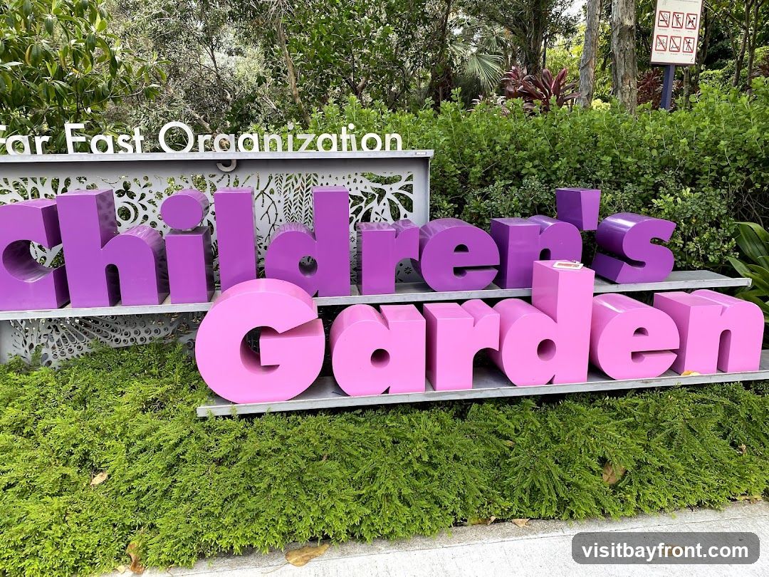 A sign for the Children's Garden, sponsored by Far East Organization. The sign features large, three-dimensional letters in purple and pink, spelling out "Children's Garden." The letters are displayed on tiered shelves against a backdrop of lush greenery. In the upper right corner, a sign with pictograms indicates rules or warnings.