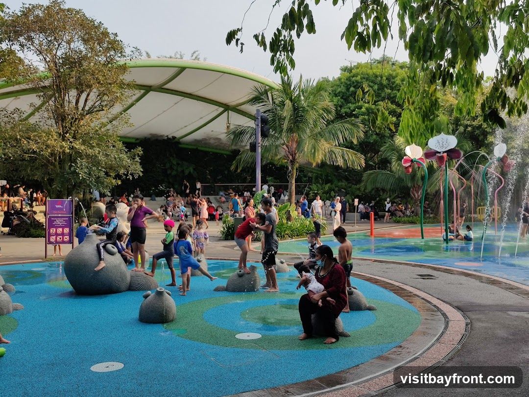 A lively water play area with children and adults enjoying the splash pads and water features. The area has a colorful, cushioned surface with various animal-shaped structures for children to climb on. In the background, there are palm trees, lush greenery, and a large, white, canopy-like structure. People are seated and standing around the perimeter, observing or resting.