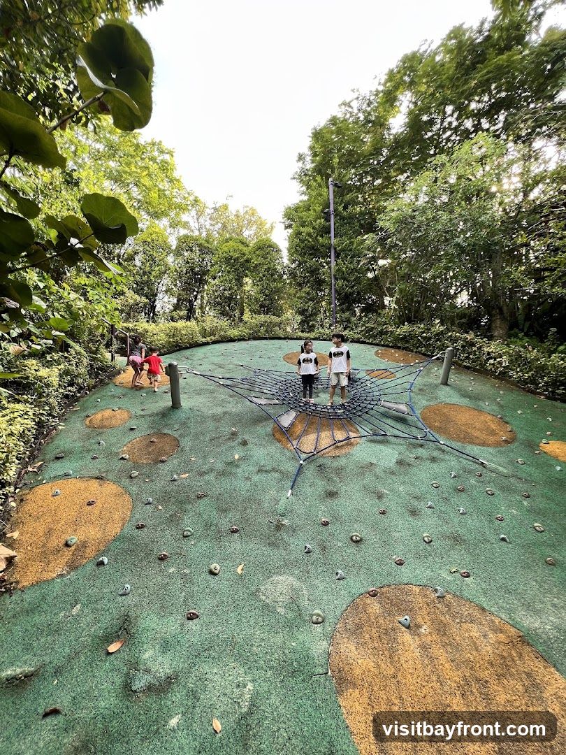 A group of children are playing on a circular playground with a net climbing structure and climbing holds. The playground is surrounded by lush green trees and foliage.
