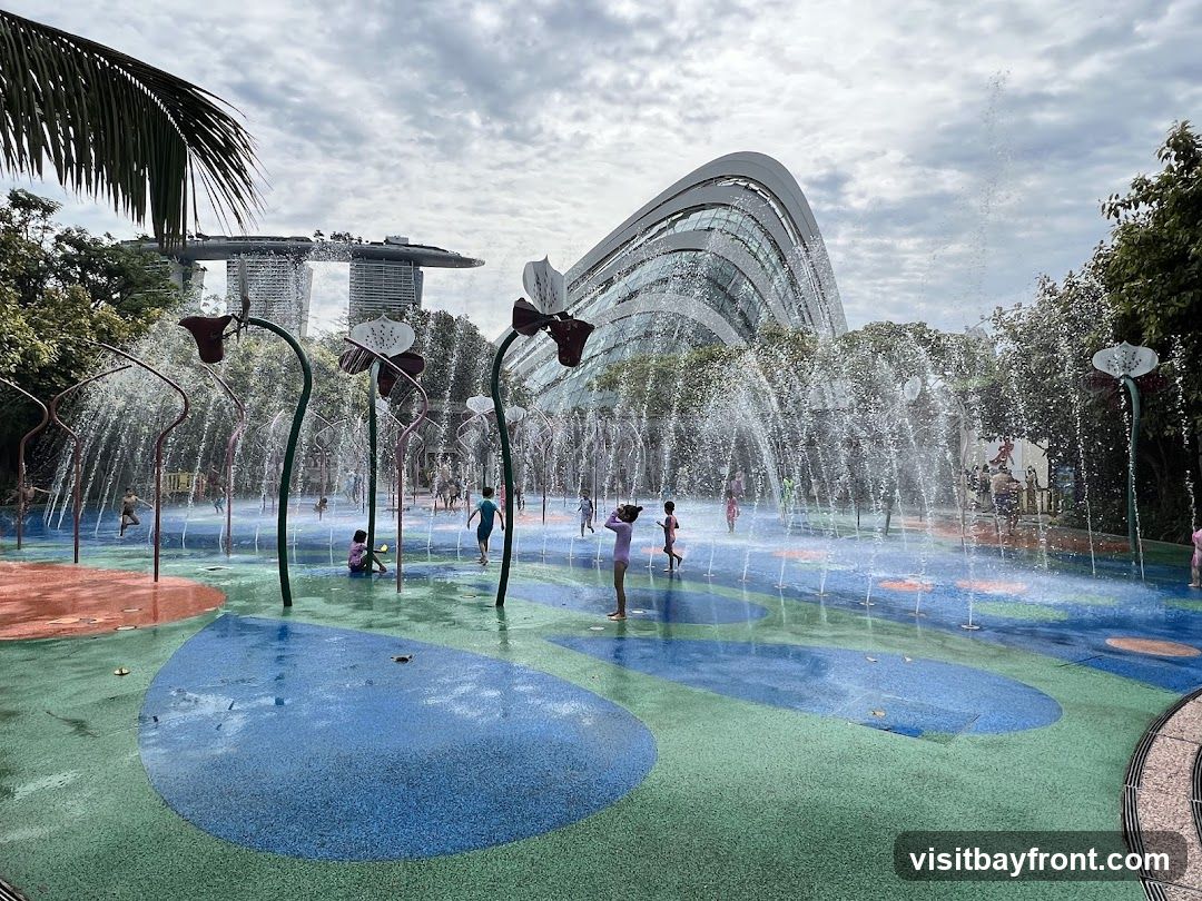 A vibrant water playground with children playing amidst sprinklers shaped like flowers. In the background, the distinctive architecture of the Marina Bay Sands hotel and the Supertree Grove at Gardens by the Bay are visible under a cloudy sky. The ground of the playground features colorful, abstract shapes in blue, green, and orange.