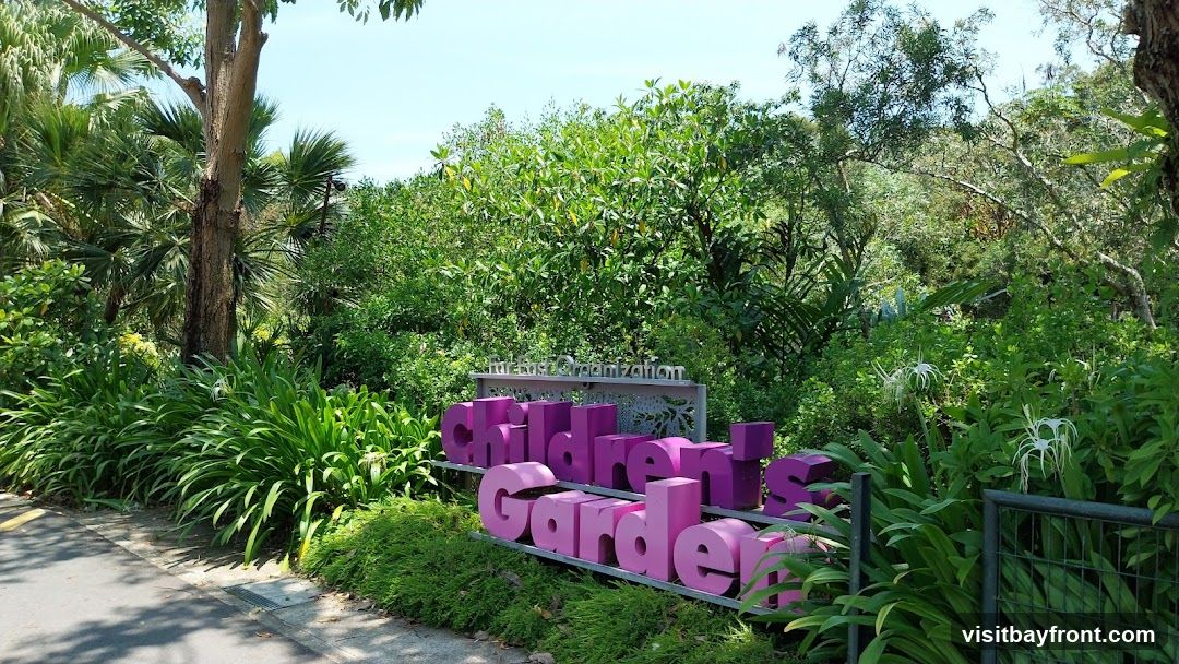 A sign for the Children's Garden is set against a backdrop of lush greenery and trees. The sign features large, purple, three-dimensional letters spelling out "Children's Garden." Above the letters, a silver sign reads "Forest Organization." The area is surrounded by dense foliage, including large-leafed plants and flowering white spider lilies.