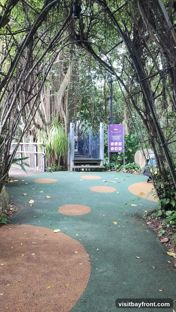 A pathway leads through a natural archway formed by intertwined branches and vines. The ground is a soft, rubberized surface with circular patterns in green and brown. In the background, a suspension bridge with wooden steps and blue railings leads upwards, surrounded by lush greenery and large trees. A purple sign with icons and text is visible to the right of the bridge.