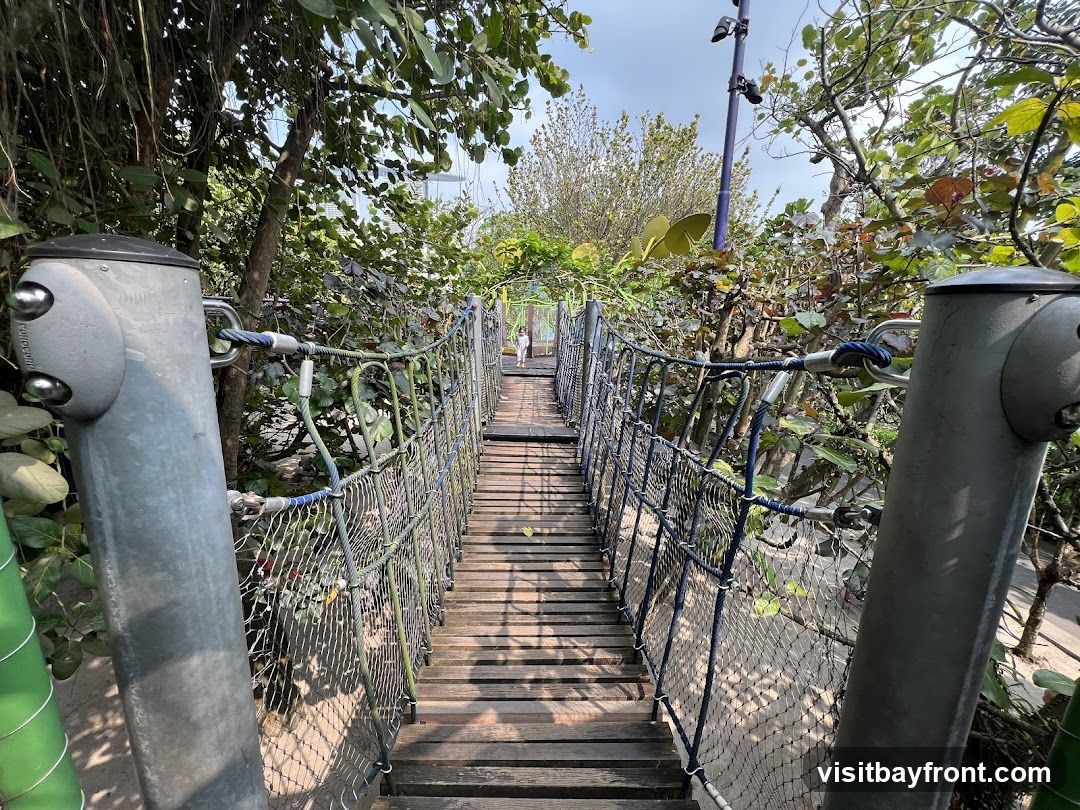 A wooden suspension bridge with rope netting on the sides winds through lush green foliage. The bridge is supported by sturdy metal posts. In the distance, a child can be seen walking on the bridge. The scene appears to be an outdoor recreational area, possibly a park or a nature attraction.