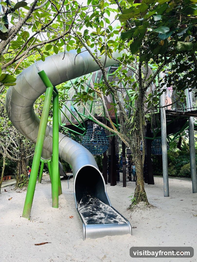 A large, winding metal slide with green support poles is set in a sandy area surrounded by lush trees and foliage. In the background, a multi-level play structure with nets and walkways is visible, with a few people exploring it.