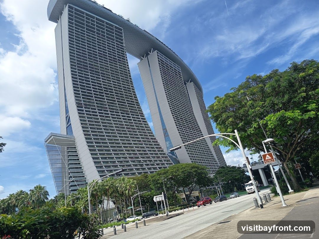 A wide-angle shot of the Marina Bay Sands hotel in Singapore on a sunny day. The iconic three towers are visible, connected by the SkyPark at the top. Lush green trees and palm trees line the foreground, with a road and some cars in the middle ground. The sky is blue with scattered white clouds.