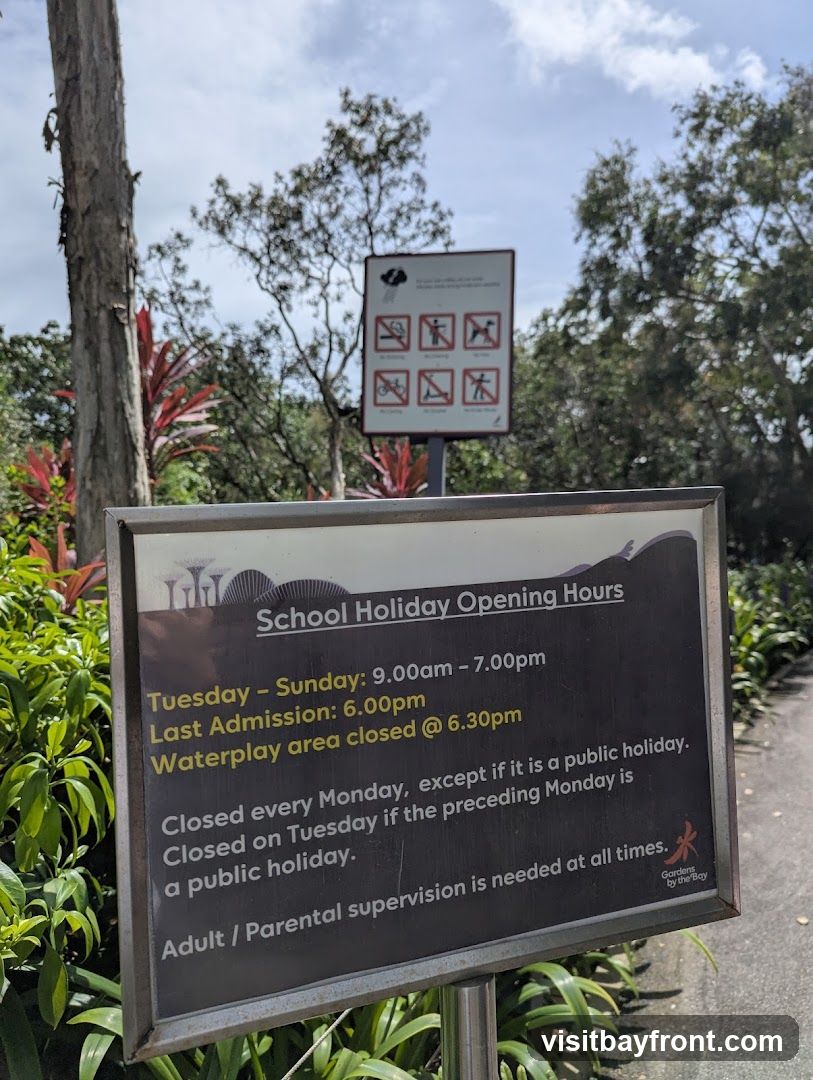 A sign displays the school holiday opening hours for Gardens by the Bay. It details the operating hours from Tuesday to Sunday, the last admission time, and when the waterplay area is closed. It also specifies closure days, including Mondays and Tuesdays if they follow a public holiday. A note about adult supervision is also present.