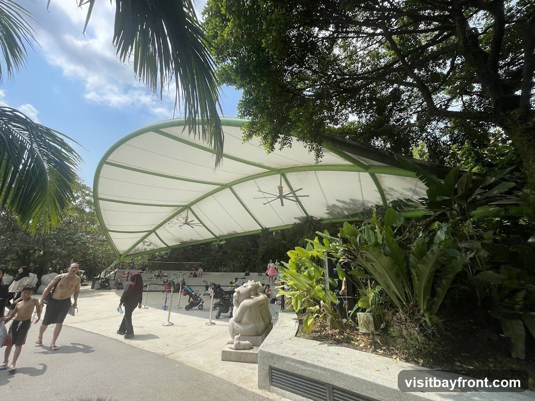 An outdoor seating area is covered by a large, white, curved canopy supported by green metal beams. Several ceiling fans are visible under the canopy. Lush green plants and trees surround the area, with palm fronds framing the top left. People are seen walking and sitting in the background, some with strollers. A stone sculpture of a creature is positioned in the foreground. The ground is paved, and the sky is blue with scattered clouds.