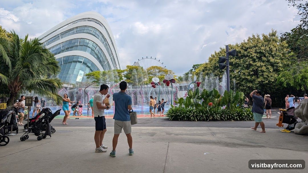 A panoramic view of a water play area with several people, including children, splashing in the water. In the background, a large, modern, curved building with glass panels is visible, along with trees and a Ferris wheel. The scene appears to be a public park or attraction on a cloudy day.
