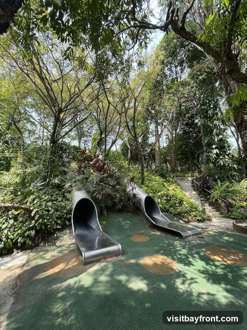 Two metal tube slides are set into a green, padded playground surface surrounded by dense foliage and trees. A pathway with steps is visible in the background, leading uphill through the greenery. The scene is bathed in sunlight, casting dappled shadows on the ground.