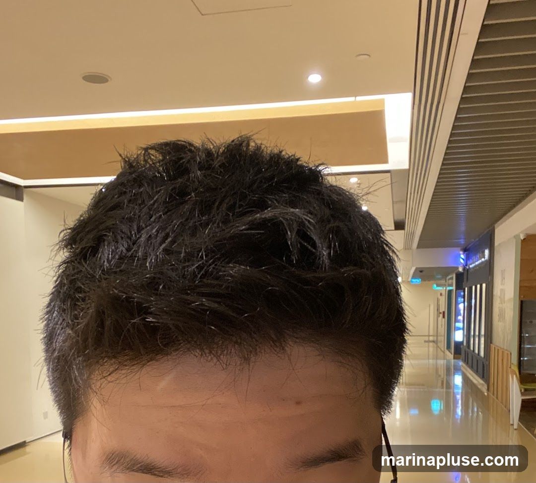 A close-up shot of a man's dark, messy hair and forehead, with a blurred background of a brightly lit indoor shopping mall corridor. The corridor features reflective floors, ceiling lights, and storefronts on the right side.