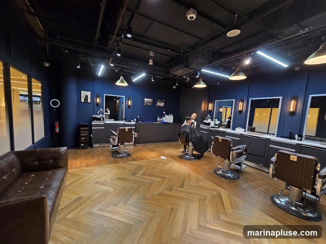 A well-lit barbershop interior with dark blue walls and a herringbone wooden floor. The shop features several barber chairs, mirrors, and a brown leather sofa. A barber is seen attending to a client in one of the chairs.