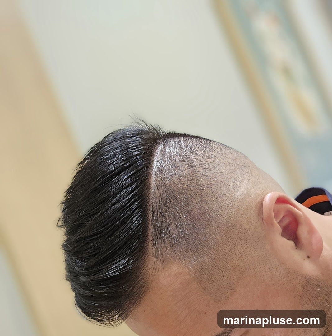 Close-up of a man's head showing a fresh haircut with a sharp side part. One side has longer, slicked-back dark hair, while the other side is closely shaved with a fade. The background is blurred and appears to be an indoor setting.